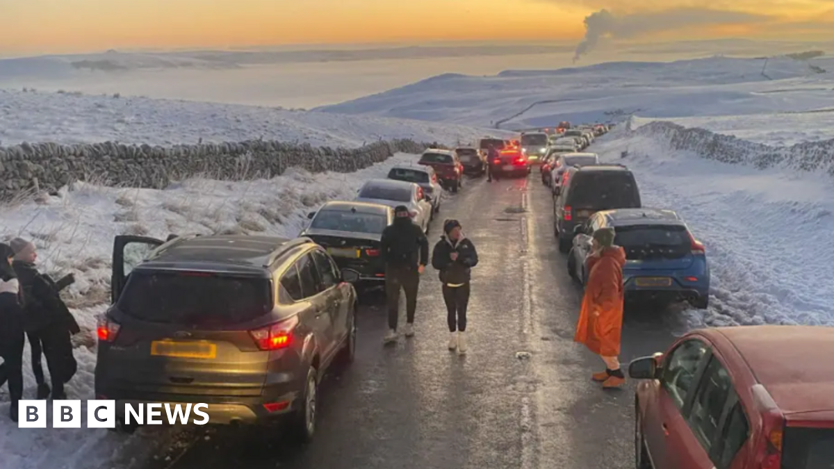 Narrow road surrounded by snowy landscape with roads parked either side of the road and some cars driving down the middle