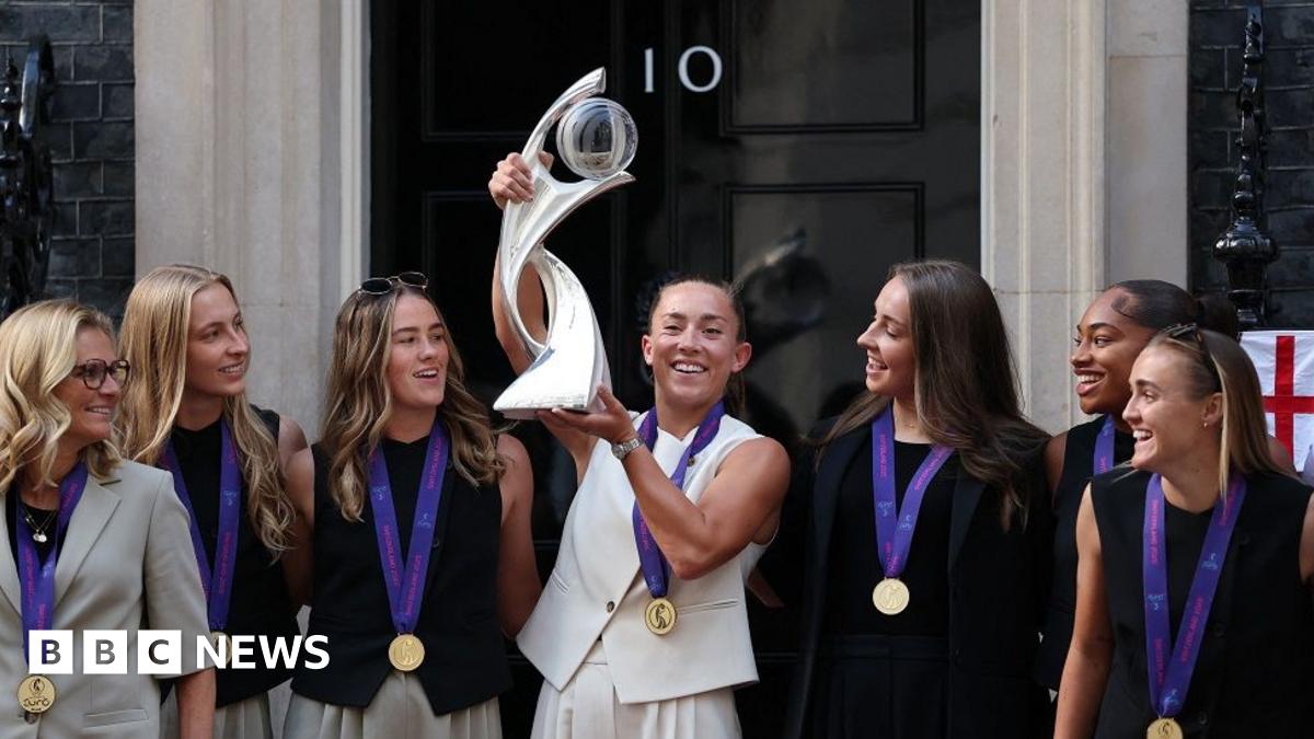 England's Maya Le Tissier lifts the trophy high above her head. Her teammates are wearing medals and smiling. They are standing in front of the No 10 Downing Street door
