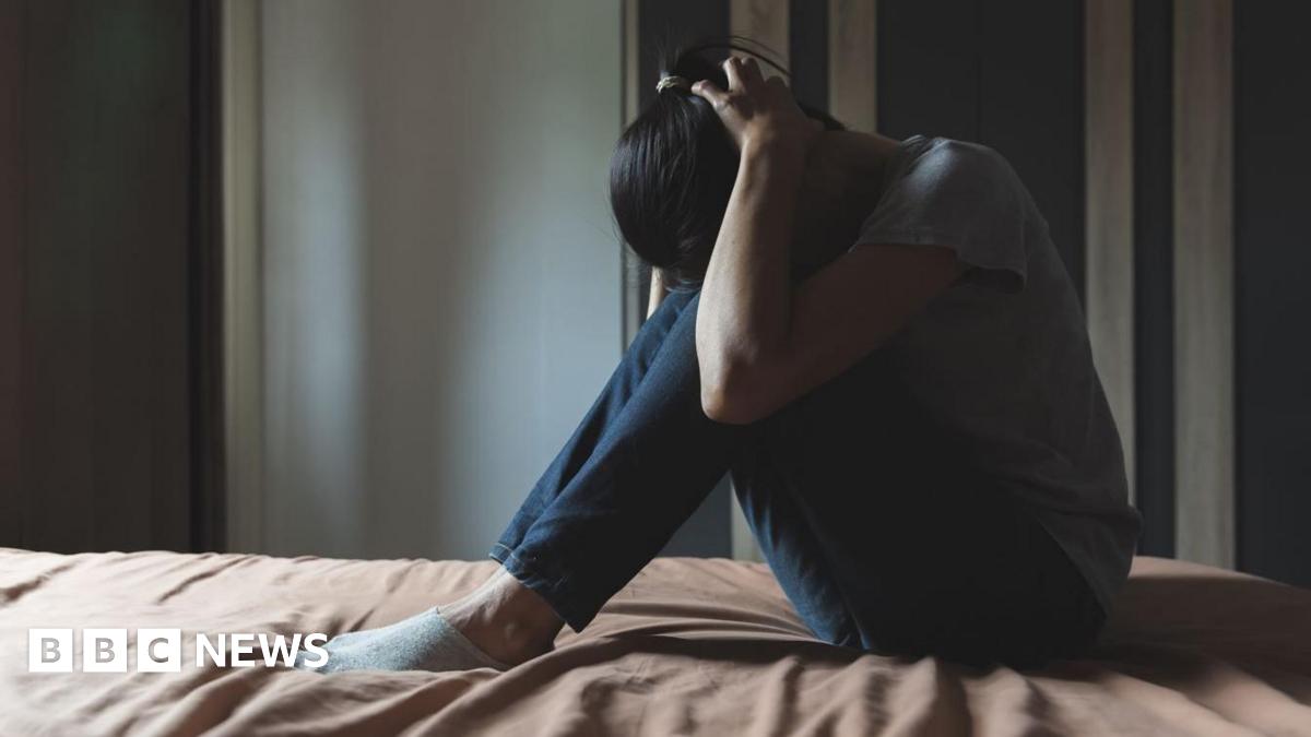 Anonymised generic photo of a woman sitting on a bed in her bedroom with her head in her hands. She is wearing a grey t-shirt and blue jeans and light coloured trainer socks and the bed sheet is light brown. 