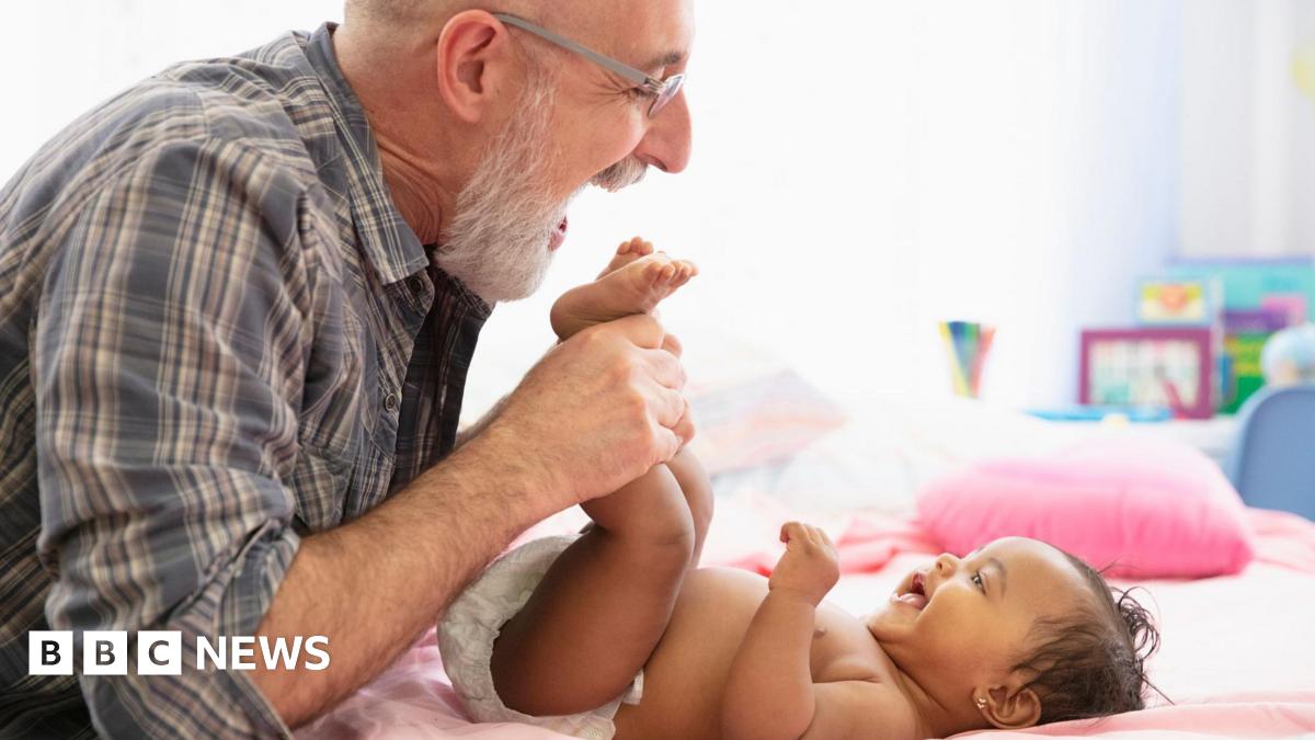 An older dad with a grey beard and wearing glasses and a checked shirt, holds a young baby's feet while she lies on a bed, wearing a nappy, and smiles up at him