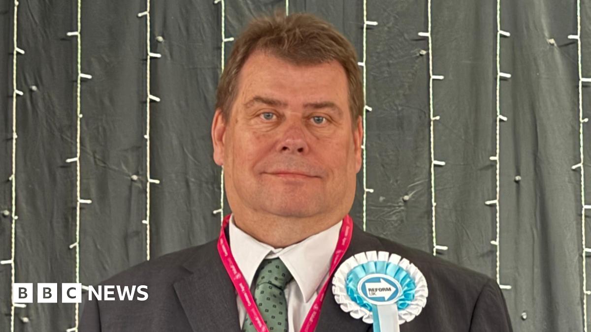 A clean-shaven man with short brown hair is weaing a black suit,  pale green dotted tie, a pink lanyard and blue and white Reform ribbon badge