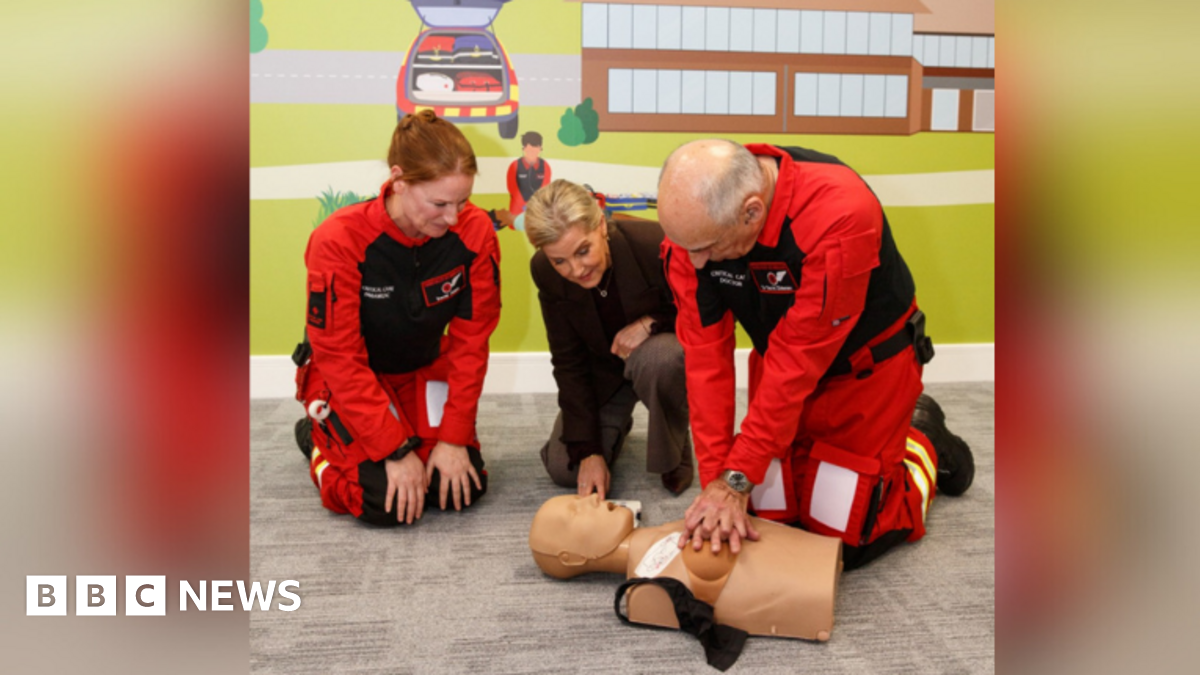 Two crew members of the Thames Valley Air Ambulance performing a CPR training on a manikin. Charity Royal Patron, The Duchess of Edinburgh, is watching them. They are kneeling in front of a colourful wall. The manikin is of a woman. The black bra acros...