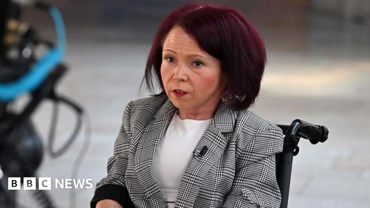 Pam Duncan-Glancy seated in a wheelchair wearing a checked blazer and white top, with a camera positioned in the foreground.