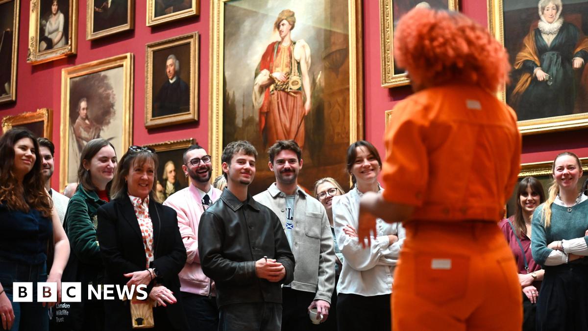 A group of people stand in the National Portrait Gallery listening to a woman in an orange outfit speak. 