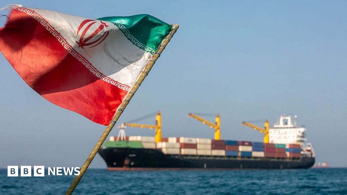 An tattered Iranian flag billowing against a blue sky with the Strait of Hormuz and a large trading ship in the background in soft focus
