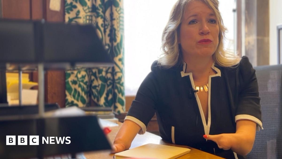 A woman with blonde hair and wearing a dark dress is sat in an office chair in front of a desk. She has foreshortened arms with only one digit on each hand. A notepad is on the desk in front of her and a stack of office letter trays. Behind her there i...