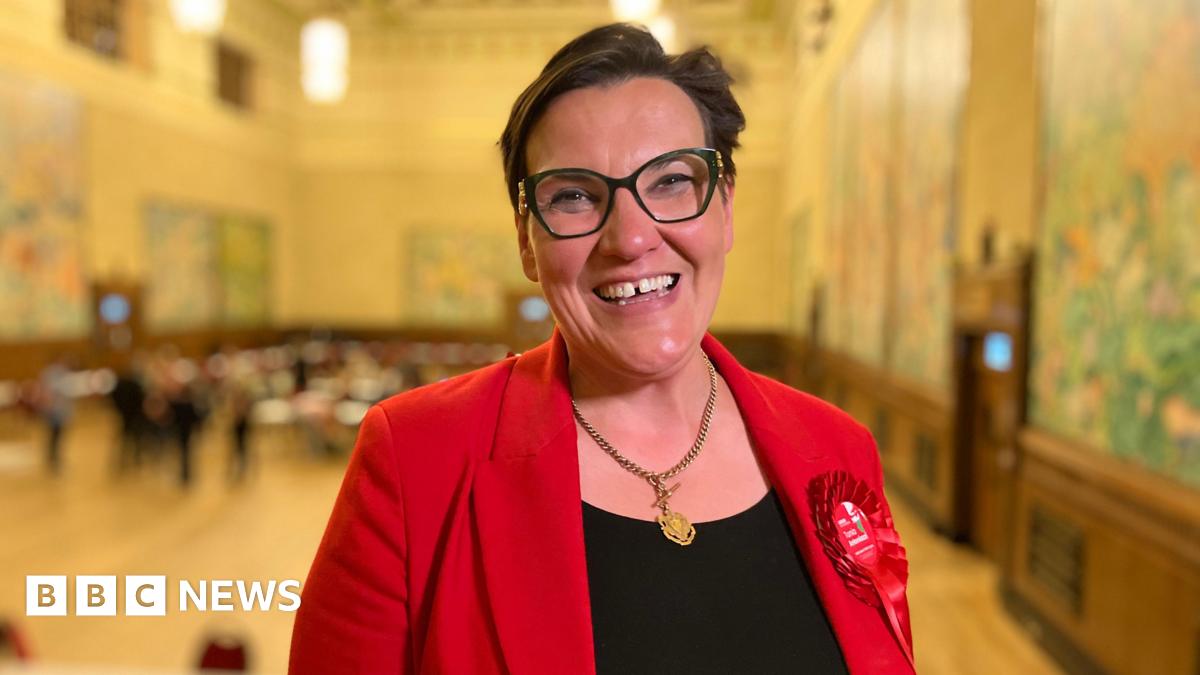Tonia Antoniazzi wearing her Labour rosette in a red blazer with a gold necklace standing in a large, ornate hall with chandeliers, murals on the walls, and people gathered around tables.