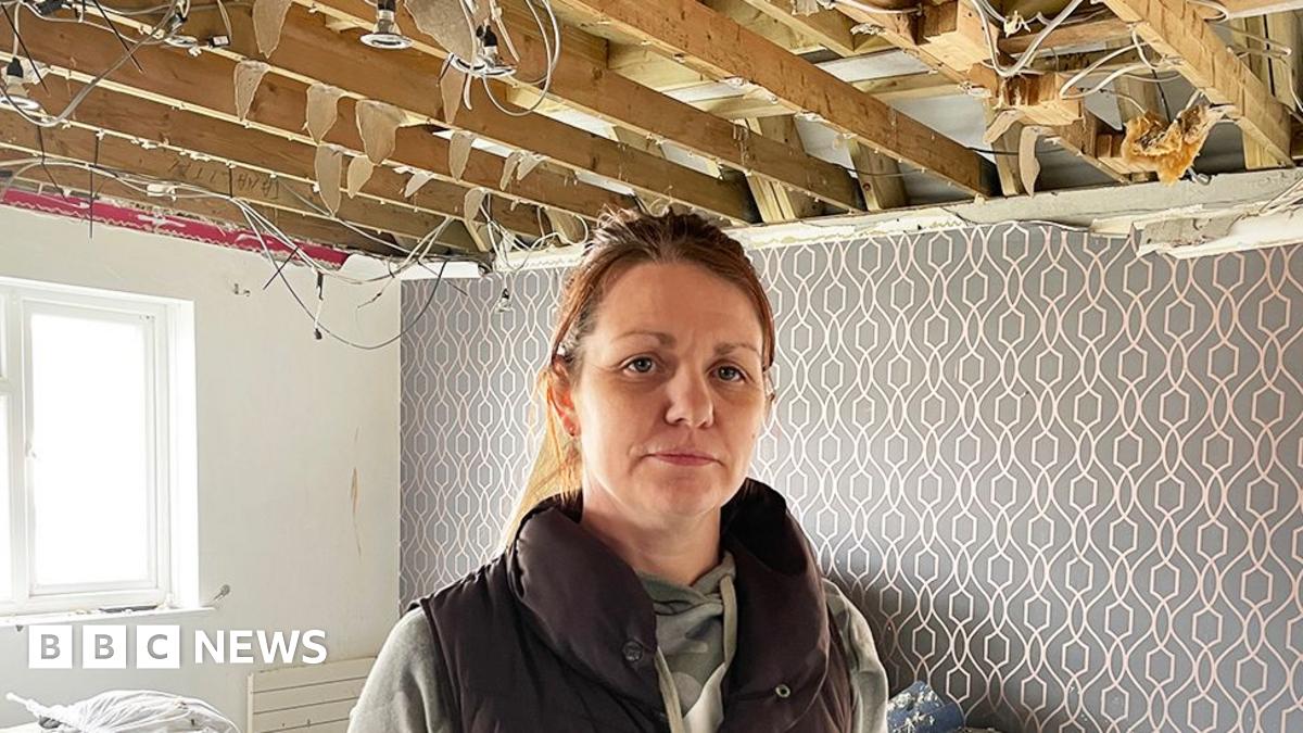 Kate Morris stands inside a severely damaged room with exposed wooden ceiling beams and hanging electrical wires. The floor is covered in debris, and the walls feature patterned wallpaper with a geometric design. A window on the left lets in natural li...