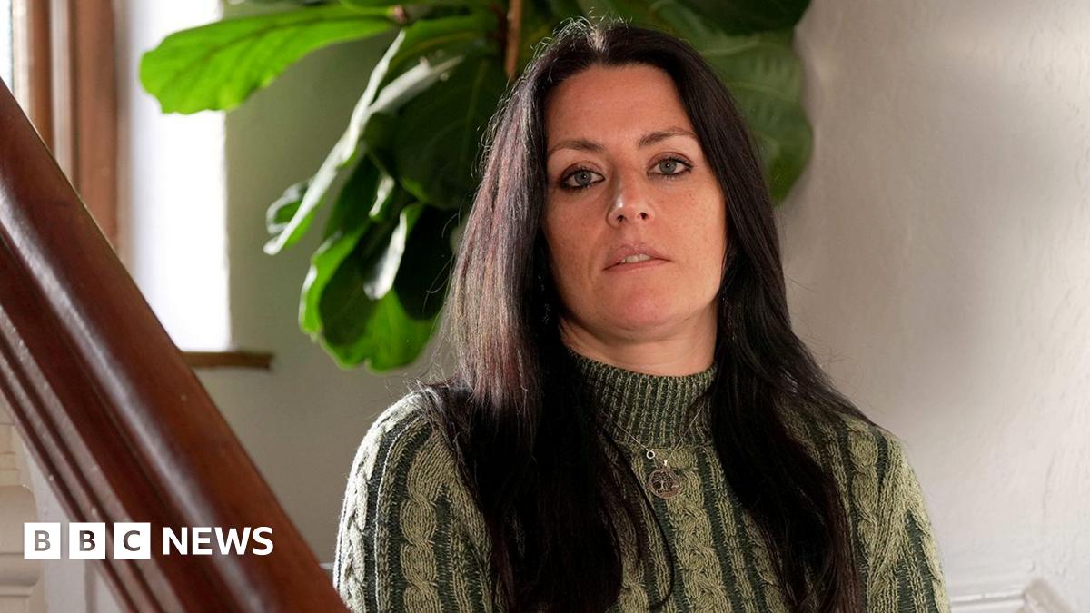 Rebecca has long dark hair and is wearing a green and cream polo neck sweater. She is sitting on the stairs with her hands on her lap and looking over the bannister into the camera. There is a plant with big green leaves behind her. She is not smiling.