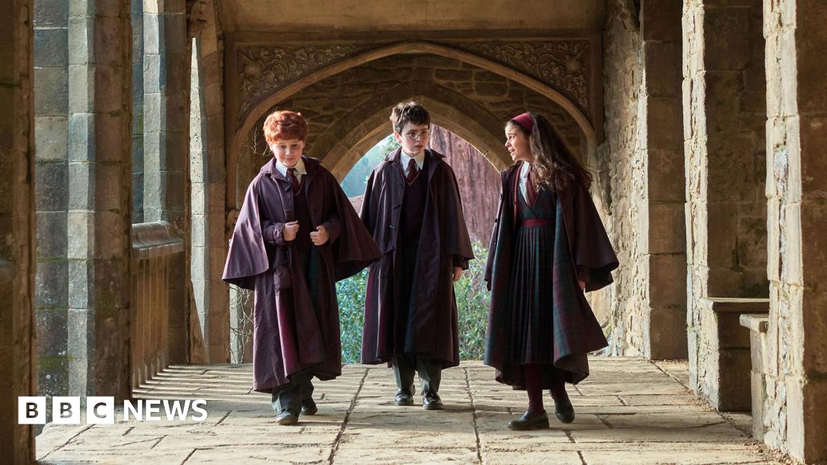 Alastair Stout as Ron Weasley, Dominic McLaughlin as Harry Potter and Arabella Stanton as Hermione Granger in the HBO YV series walking along a cloister of fictional Hogwarts school set. 