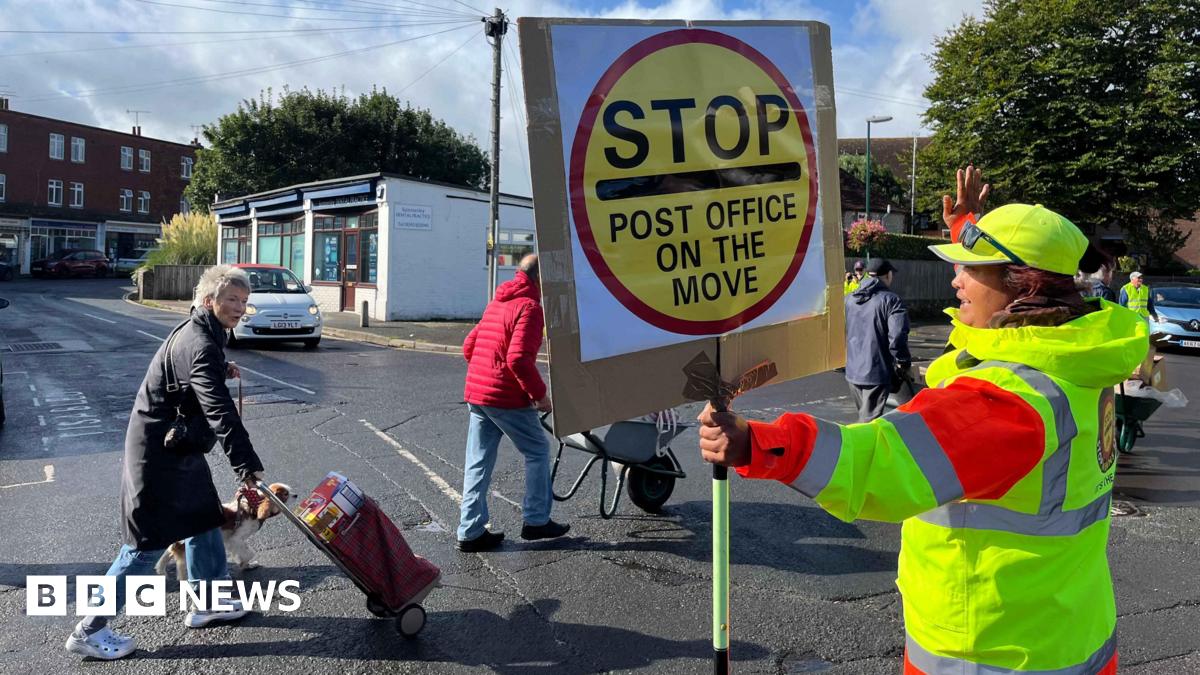 A lollipop lady in the road with a sign saying stop post office on the move. In front of her crossing the road are people with granny bags and wheel barrows. A car is waiting behind