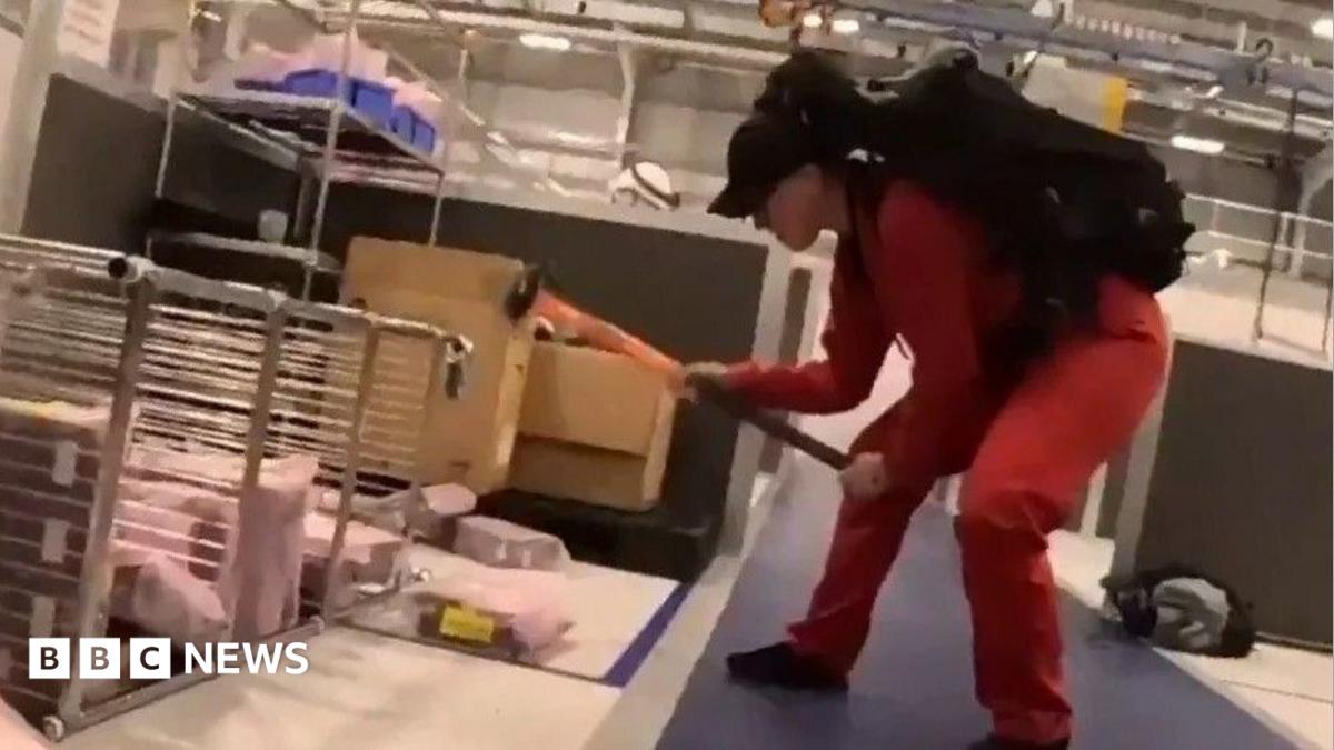 A person wearing a red jumpsuit and black cap, using a sledgehammer to damage equipment inside the factory.