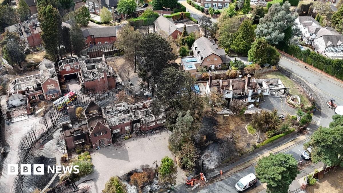 Aerial view of a large residential property and its grounds heavily damaged by fire, with emergency vehicles present on the street.