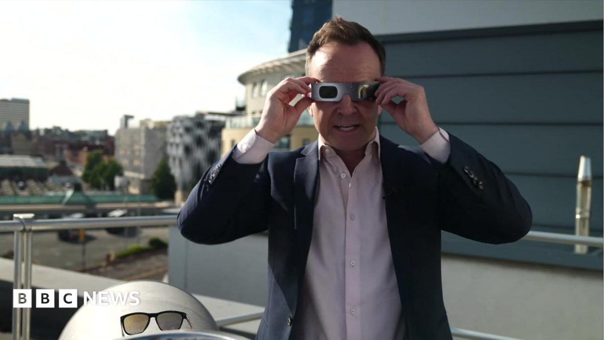 A man in a blue suit and pink shirt holds up solar eclipse glasses