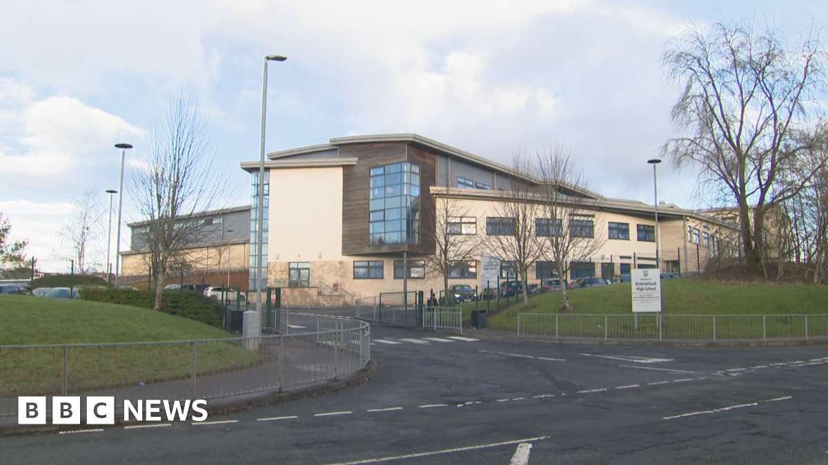 A view from across the street of Kirkintilloch High School - a large sign with the school name on it is sitting on grass, while a road leads into the premises. The building is mostly tan and brown shades, with a car park nearby. 