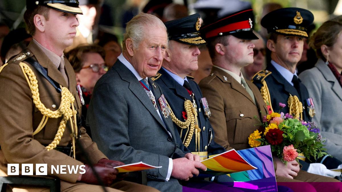 King Charles looks on while holding a ceremony programme booklet that has the LGBT+ rainbow flag colours on its cover, he's wearing a formal suit and is sitting next to various Army officers wearing formal military attire including military hats.
