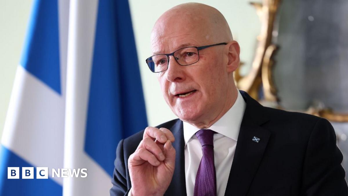 A bald man with glasses speaks to the media. He is wearing a black suit, white shirt and purple tie. He is holding his left hand in front of his chest. There is a saltire and a mirror in the background. 