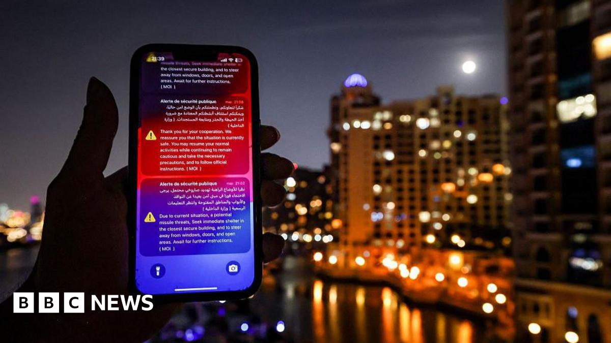Phone held up to the camera at night, on it are a stream or emergency messages. In the background are a number of high-rise buildings