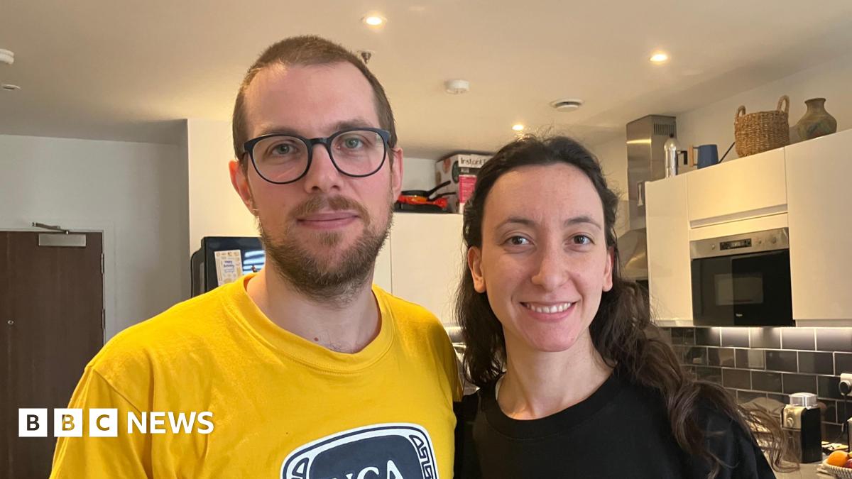 A man in a yellow top stands next to a woman in a black one. He has short brown hair and glasses. She has long dark brown hair