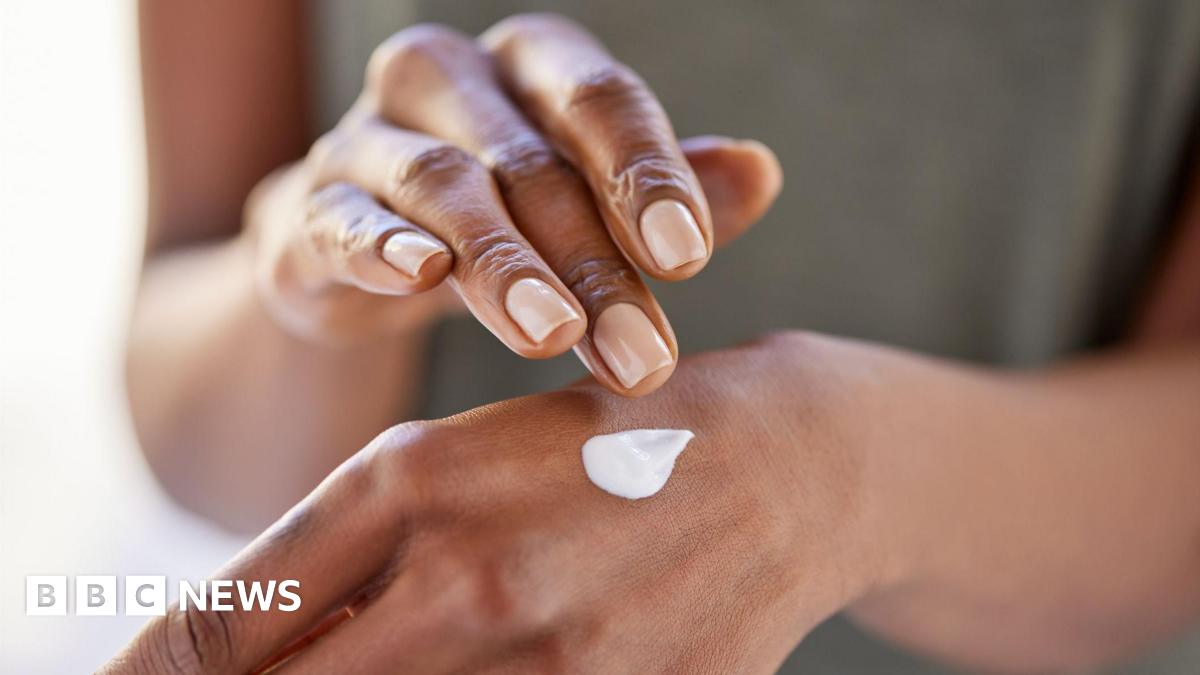 A close up of a black woman applying a white cream to her left hand with her right hand.