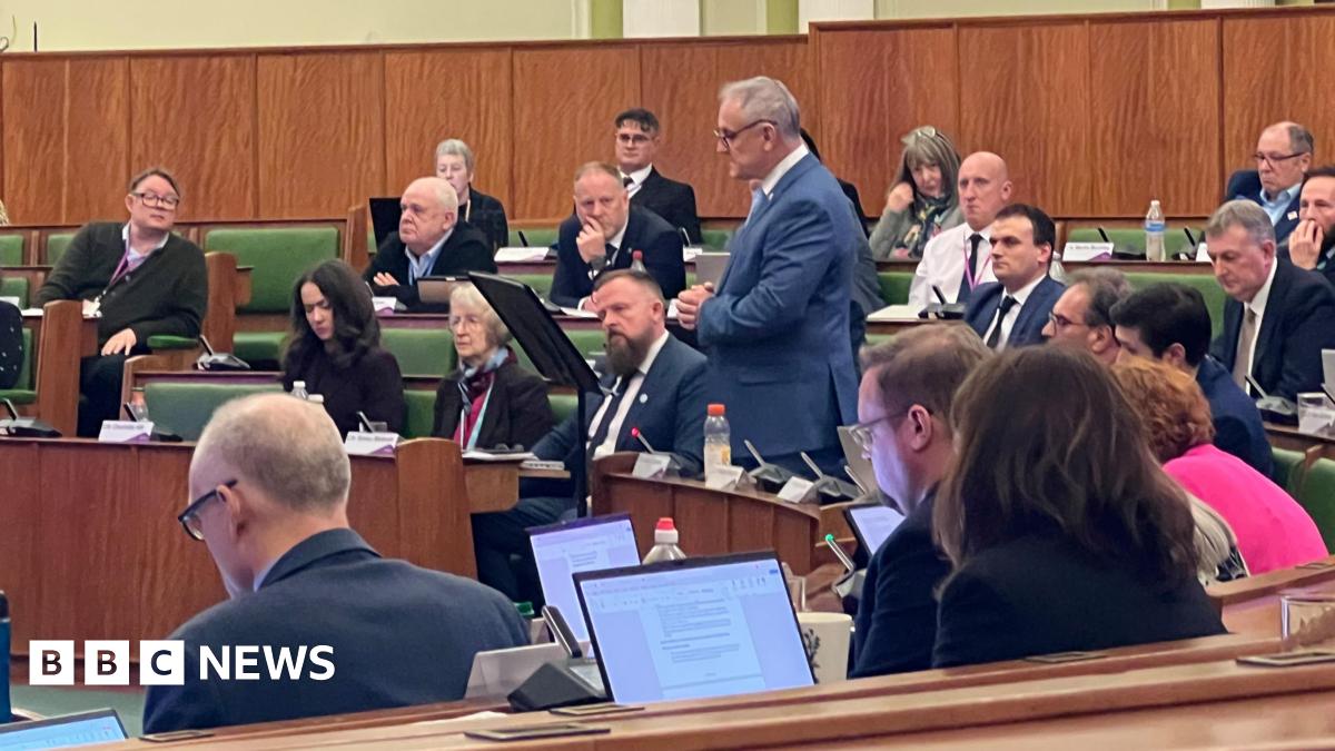 A man stood making a speech in a council house with rows of chairs full of people