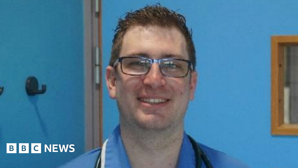 A man in a nursing uniform smiles at the camera. His uniform is blue and there is a stethescope around his neck. He has short brown hair and wears rectangular spectacles. The room his is standing in is painted bright blue.