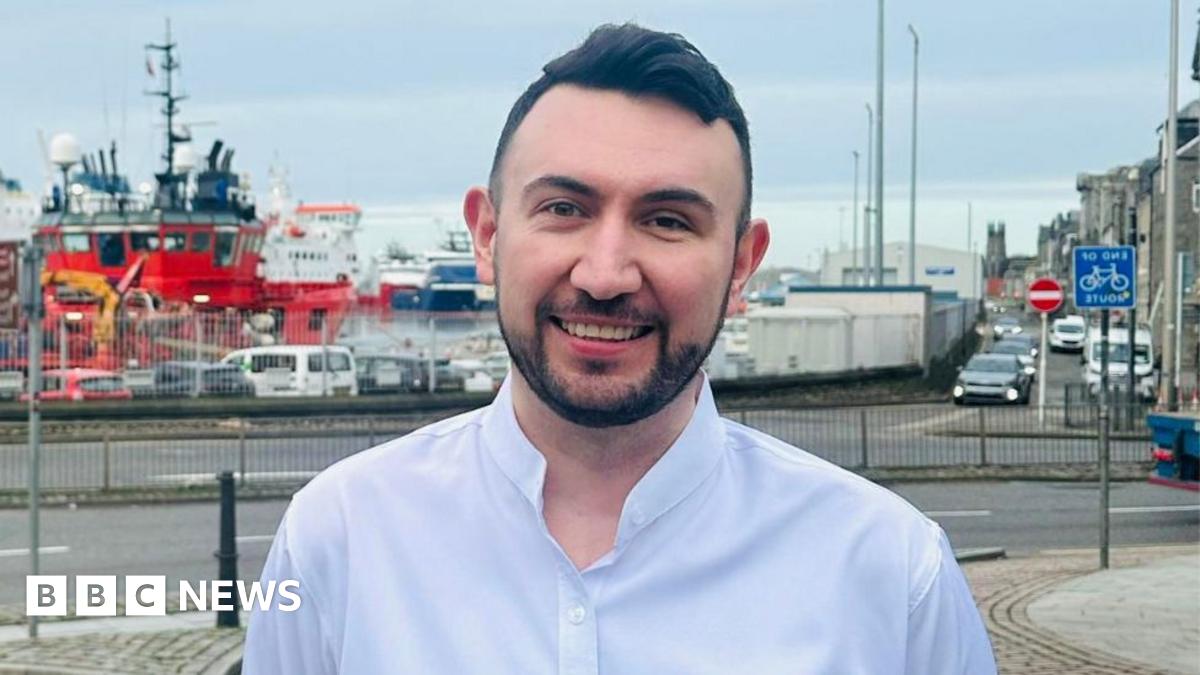 A man with dark hair and a dark beard smiles at the camera while standing in front of a harbour 