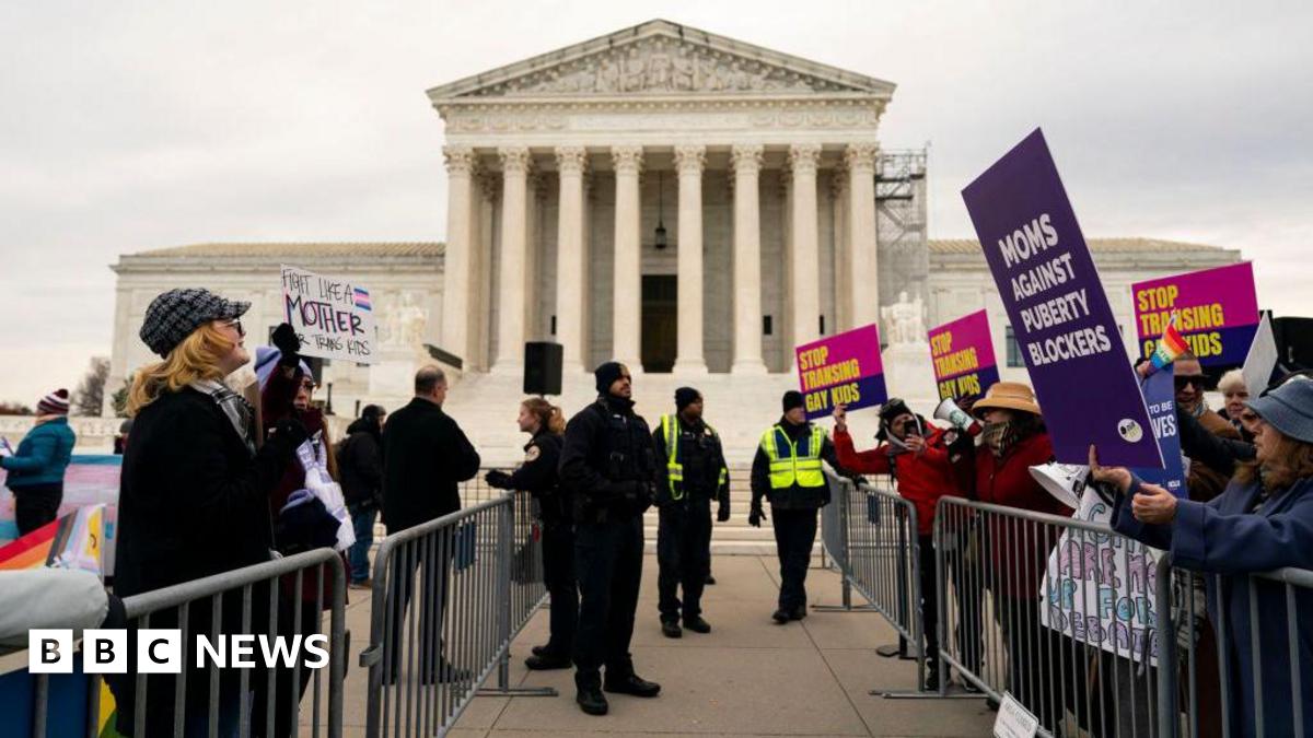 Groups of protestors stand on either side of crowd control barriers outside the Supreme Court building in Washington DC. People hold signs in support of and against gender transition treatment for children. Uniformed police officers stand in an aisle b...