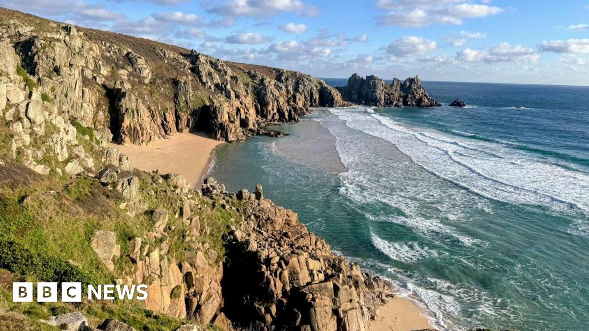 Overlooking Pedn Vounder beach. A rocky cliff face leads down to a sandy beach. Waves are rolling onto the shore. The sky is cloudy.