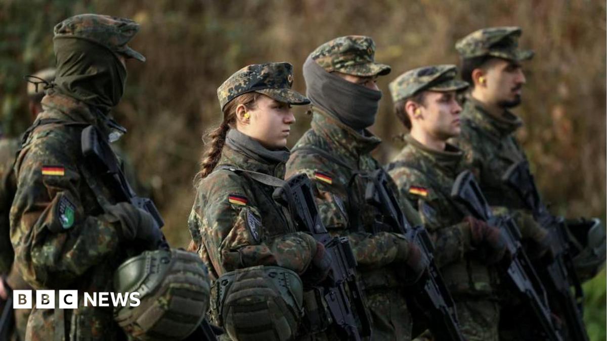 Male and female soldiers stand with guns in a field, wearing camouflage with the German flag stitched on their upper arms.
