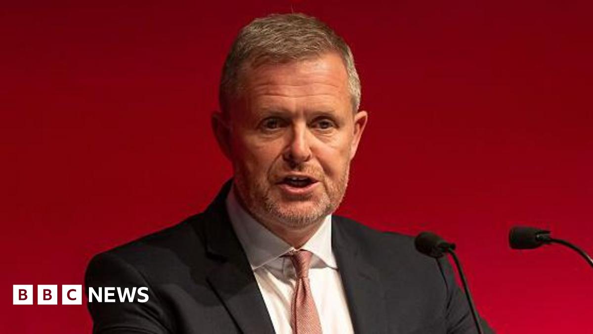 Jeremy Miles, who has grey/light brown hair and a beard wearing a black suit with a thin red lie tie over a white shirt, speaking into two small black microphones in front of a red background