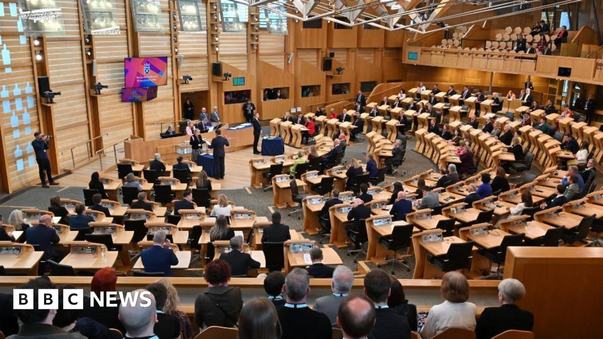 A wide shot of the Scottish Parliament chamber, with MSPs sitting at wooden desks in a wood-panelled room 