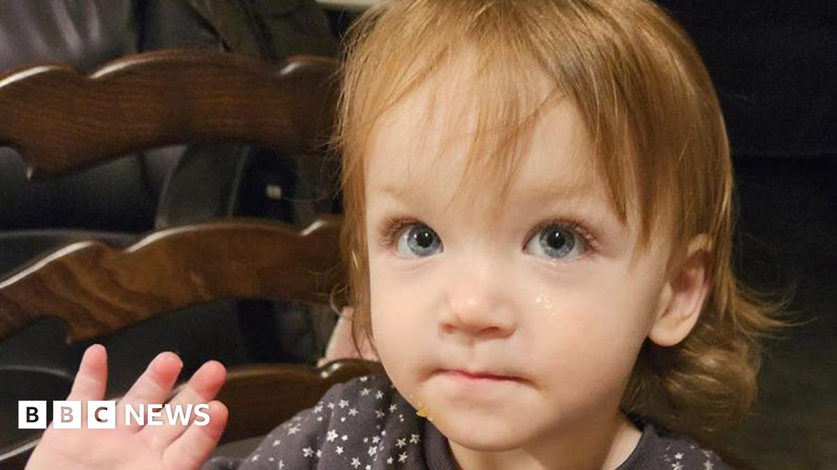 A two-year-old girl with shoulder-length brown hair and blue eyes is wearing a dark top with white stars on it. She is sitting on a chair and is holding up her right hand