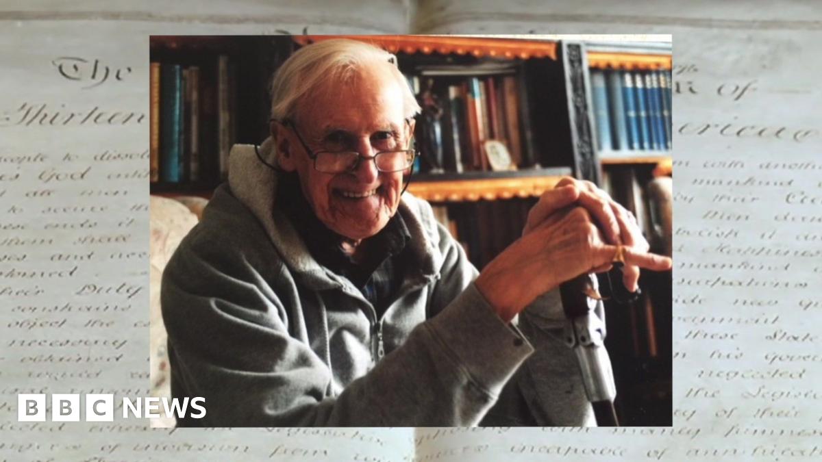A smiling elderly man in glasses with a walking stick sits in front of shelves stacked with old books