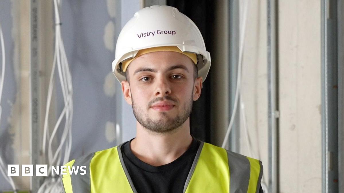 Will Cunningham, a man in a white hard hat and hi-viz tabard, looks at the camera. He has a brown thin beard and moustache