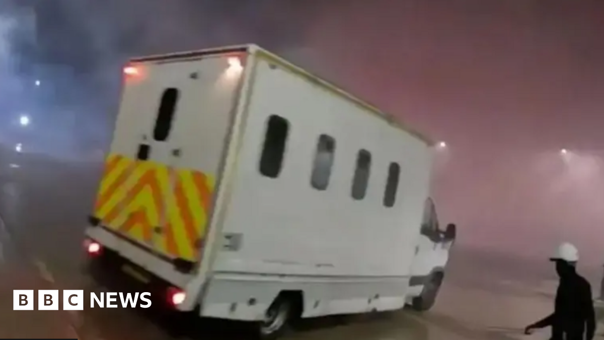 A screenshot of the police van seen on the Elbit Systems UK site. The van is a large white vehicle with small windows and a chevron pattern across the back. A figure wearing black with a white helmet can be seen in the lower right corner of the image h...