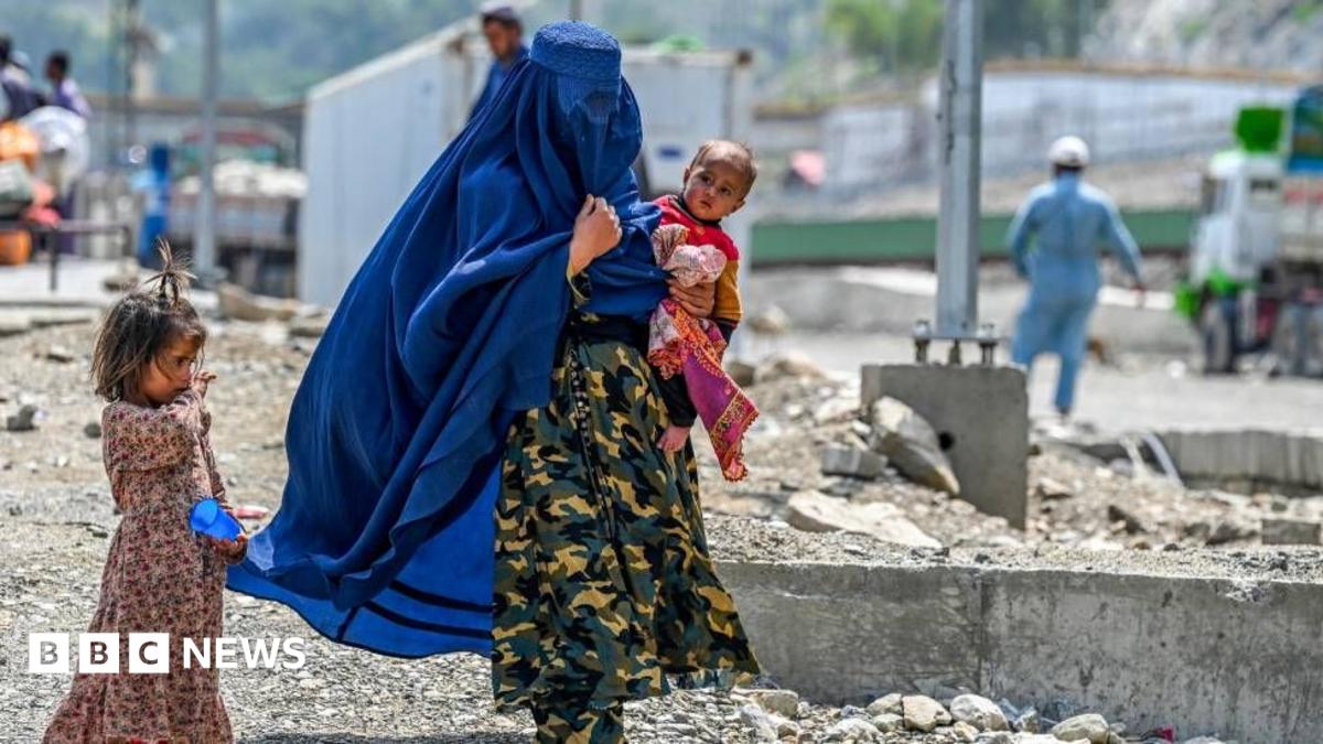 A woman carries a child in her arm. A young girl is walking alongside her. The woman is wearing an outfit in camouflage colours, and her face is covered with a long, blue veil