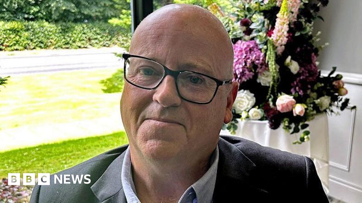 Mark McCollum wearing a blue shirt, blazer and glasses. He is looking at the camera.