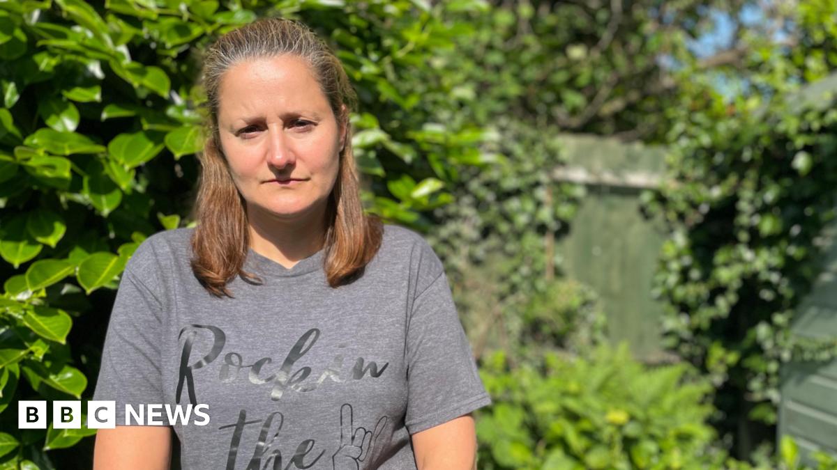 Emily Fryer, with tied-back brown hair and a dark-blue t-shirt, stands in front of a green bush in her garden and is looking solemnly at the camera