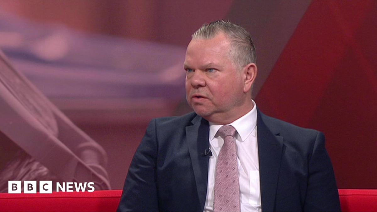 A man wearing a suit and pink tie sits on red sofa looking at the camera. A screen can be seen behind him.