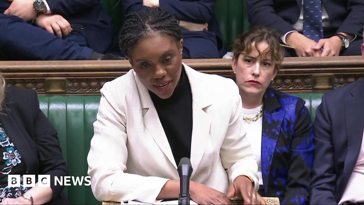 Conservative Party leader Kemi Badenoch stands at a microphone in the House of Commons. She wears a white suit jacket and black shirt.