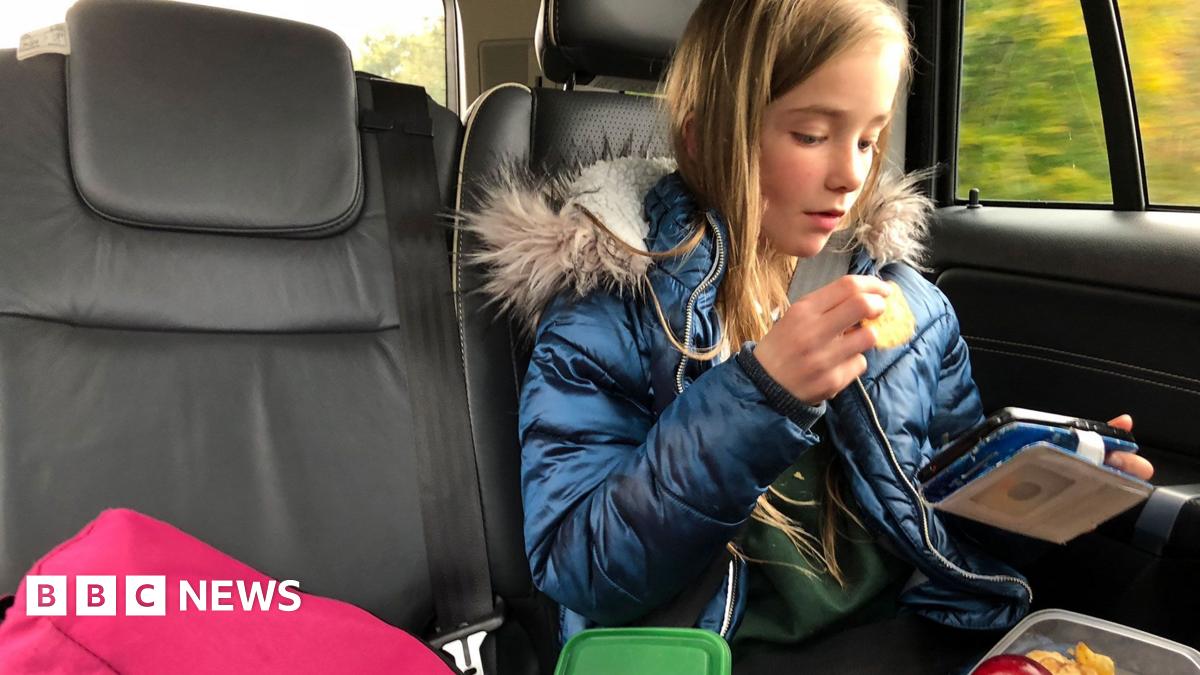 A child sits in the back on a car looking at a hand held electronic device and eating a snack from their lunch box. She is wearing a school uniform and winter coat