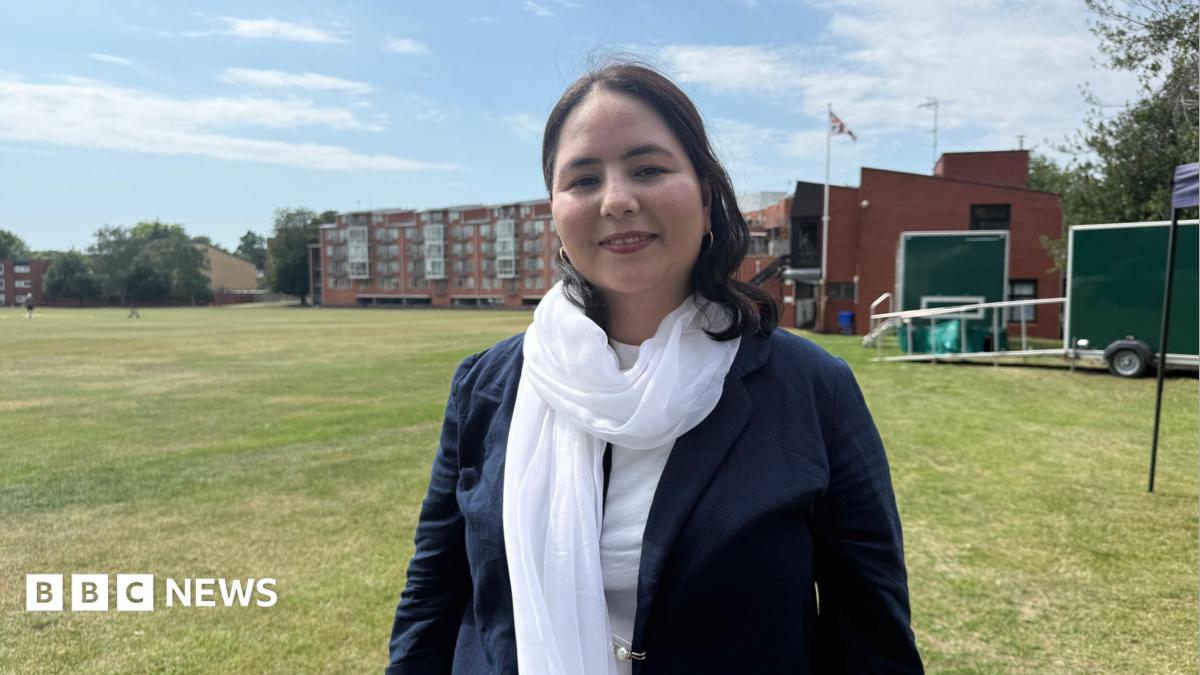 A woman with long dark brown hair wearing a blue jacket, a white scarf and a white top. She is standing on a grass sports field.