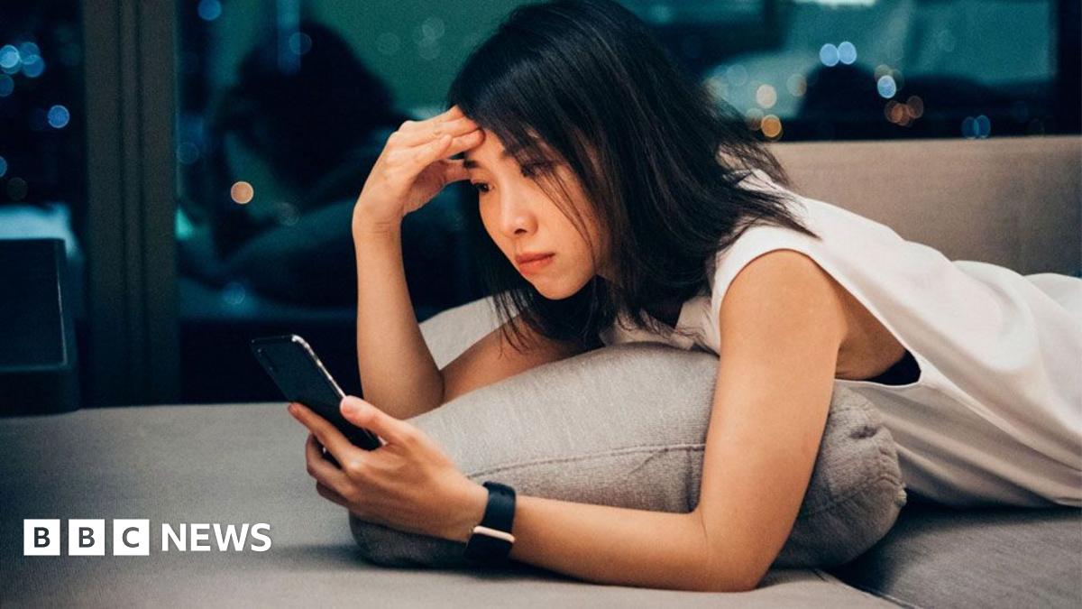 A woman lies on her front on a lounge, holding a phone in one hand and pressing her brow with the other
