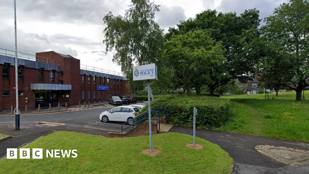 The outside of Pudsey Police Station. A red brick building is on the left of the picture with cars parked opposite. On the right are trees and a grassed area 