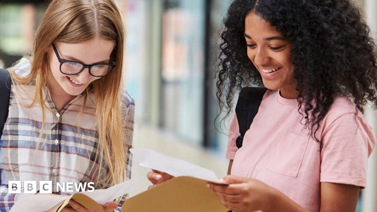 Two female students smile as they look down at exam results they've just removed from brown envelopes. The student on the left has long straight blonde hair, and wears thick-rimmed black glasses and a blue, white and red checkered shirt. The student on...