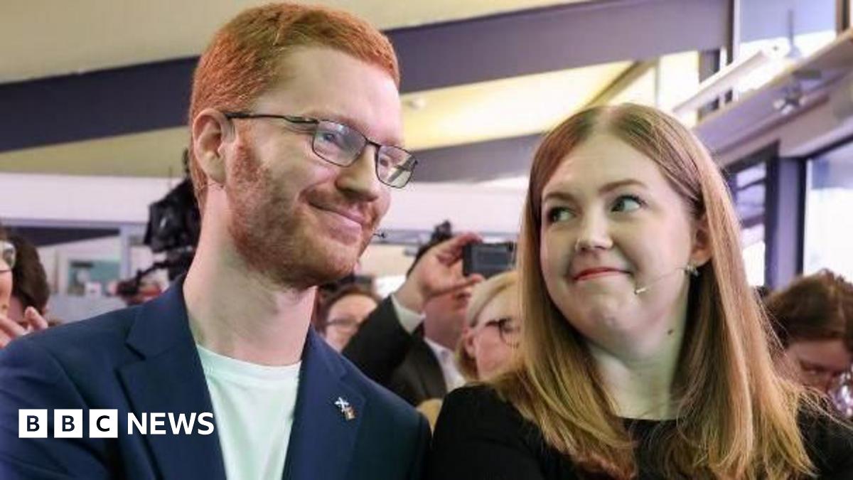 Ross Greer, who has ginger hair and glasses, and Gillian Mackay, who has fair hair and is wearing a black jumper, sit next to each other in the front row of an audience. Greer, wearing a navy suit jacket over a white t-shirt, has his right hand on Mack...