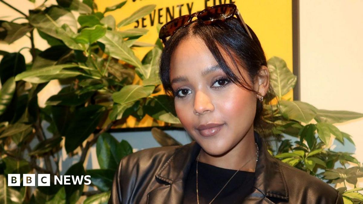 Winta Zesu wearing a black leather jacket stands in front of plant.