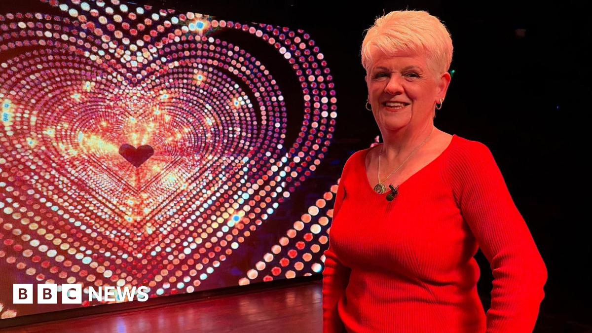 Sheila Diamond is standing on stage in front of a screen with a sparkly heart. She is wearing a red, long-sleeved top and is smiling at the camera. 