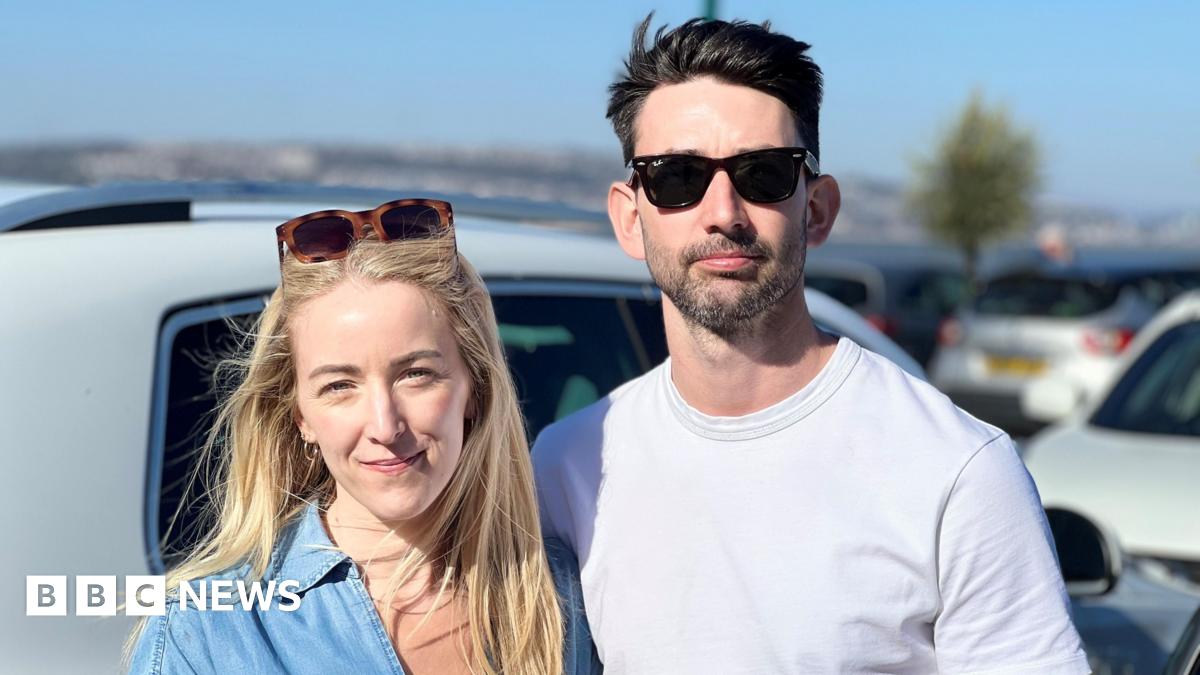 Josh and Georgina stand arm in arm in a car park. she has long blonde hair and a denim shirt, he has short cropped dark hair and a white round neck shirt. 